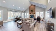 Located on Brigstock Court in Fredericksburg, Virginia, this single-story clubhouse offers a well-appointed communal space designed to serve as both a welcoming gathering area and a functional leasing office. The architecture is defined by dramatically vaulted ceilings that rise to a sharp peak, amplifying the sense of openness throughout the main area. Light wood-tone flooring runs continuously underfoot, complementing the neutral gray wall palette and white trim details.

The centerpiece of the main lounge area is a full-height brick fireplace, extending from floor to ceiling peak, flanked by large white-framed windows that draw in generous natural light and offer views of the surrounding landscape. Comfortable seating arrangements, including a sectional sofa and accent chairs, are thoughtfully positioned around the fireplace, creating an inviting conversational space. Nearby, a round table with leather chairs provides an additional seating option.

A dedicated workspace area features a curved desk with guest seating and expansive bay windows overlooking an outdoor pool and courtyard area. A secondary workstation with multiple monitors is positioned along another windowed wall.

A compact kitchenette includes dark cabinetry, stone-look countertops, a sink, and a breakfast bar with bar seating. Recessed lighting throughout ensures consistent illumination, enhancing the bright, professional ambiance of the overall space.