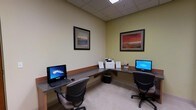 This single-floor business center is thoughtfully designed to support focused, productive work in a clean and comfortable environment. The room features an L-shaped granite-topped workstation that wraps along two walls, providing ample desk space for multiple users simultaneously. Two desktop computers are conveniently positioned at either end of the workstation, accompanied by ergonomic office chairs that ensure comfort during extended use. A multifunction printer sits centrally along the desk, making printing, scanning, and copying easily accessible. The drop ceiling is fitted with recessed panel lighting that evenly illuminates the workspace, creating a bright and professional atmosphere. Light-colored walls with a warm undertone and a wood-trimmed wainscoting accent lend a polished, welcoming feel to the room. Two framed landscape artworks hang on the walls, adding a subtle decorative touch that softens the functional setting. The neutral carpeted flooring completes the space with a quiet, professional finish. Overall, this business center is well-equipped and efficiently laid out, offering everything needed for productivity in a tidy, distraction-free setting.