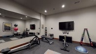 This single-floor recreational facility is thoughtfully divided into distinct activity zones, offering a comprehensive range of fitness and entertainment options. The fitness room is equipped with professional-grade exercise equipment, including a Matrix rowing machine, a stationary cycling bike, a weight bench, and a medicine ball rack. Rubber flooring provides a safe workout surface, while recessed lighting and a large wall mirror enhance the functional atmosphere. A wall-mounted flat-screen television adds to the convenience of the space.

The adjoining game room is generously proportioned and features dark hardwood-style flooring and neutral walls accented by warm wood baseboards. Large windows allow natural light to fill the room, offering views of the surrounding neighborhood. A full-size pool table with a blue felt surface serves as a centerpiece, complemented by a ping-pong table, a wall-mounted dartboard cabinet, and a rustic wood high-top table with bar stools. Additional wall-mounted televisions are positioned throughout for viewing convenience. Framed wildlife and nature artwork adds a distinctive decorative touch. Recessed and pendant lighting complete the inviting, activity-focused ambiance of this well-appointed space.