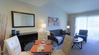 This single-floor residence showcases a thoughtfully designed open-concept layout that seamlessly integrates the dining and living areas into one cohesive space. A standout architectural feature is the vaulted ceiling, which rises to a peak and adds a sense of airiness and volume to the room.

The living area is anchored by a dark blue sofa accented with decorative pillows, complemented by warm ambient lighting from floor lamps on either side. A colorful abstract canvas adds a vibrant focal point to the neutral gray walls. Natural light filters in through a window, brightening the space and enhancing its welcoming atmosphere.

The dining area features a round glass-top table paired with tufted cream chairs, offering a modern and elegant touch. A dark wood media console with a large framed mirror sits along one wall, adding depth and style.

Neutral-toned carpet runs throughout, contributing to the space's warm and comfortable feel. The overall ambiance is clean, modern, and well-appointed, balancing functionality with tasteful décor throughout.