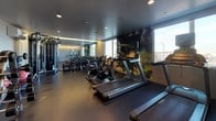 This single-floor fitness center at 3004–3010 San Pablo Ave in Berkeley, California, offers a thoughtfully designed workout space with a modern, functional aesthetic. The interior features dark rubberized flooring throughout, recessed ceiling lighting, and a neutral color palette that gives the space a sleek, professional atmosphere.

The main workout area is equipped with multiple treadmills, Stages cycling bikes, and elliptical machines arranged in a row along one wall, with a large window providing natural light and outdoor views. A decorative nature-themed wall mural adds a visual focal point alongside the cardio equipment. Ambient underlighting along the upper walls contributes to the contemporary feel of the space.

A dedicated functional training zone houses a multi-station cable machine system with TRX suspension straps, along with stability balls, medicine balls, a BOSU trainer, and a rack of free weights, offering a full range of strength and conditioning options. A glass partition separates distinct zones within the facility.

Near the entrance, practical amenities include a paper towel dispenser, a water fountain, and a waste bin, ensuring a clean and hygienic environment. Signage at the entry door outlines facility guidelines, including a no-pets and no-smoking policy.