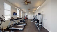 This fitness center is housed within a single-story space designed to accommodate a full range of exercise activities. The layout is elongated and open, allowing equipment to be organized into clearly defined zones without feeling crowded. The flooring throughout is durable rubber gym matting, providing traction and impact absorption suitable for both cardio and strength training use.

Cardio equipment is prominently featured, including multiple treadmills, elliptical trainers, and stationary bikes arranged along one side of the room. The strength training section offers a multi-station cable machine, a functional trainer, a rack of dumbbells spanning a wide weight range, an adjustable bench, and a medicine ball rack. Stability balls and additional free weights round out the available equipment.

Natural light enters through several large windows fitted with horizontal blinds, brightening the space considerably. Multiple wall-mounted flat-screen televisions are positioned for visibility throughout the room. Ceiling fans are evenly spaced across the ceiling, supplementing the HVAC ventilation system. Linear LED lighting fixtures run along the ceiling, ensuring consistent illumination. A large wall mirror in the free weights area enhances the sense of space and supports proper form during training.
