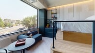 This cleverly designed studio unit maximizes every inch of its footprint through smart spatial planning and multi-functional design elements. Upon entry, a wall-mounted coat rack and framed artwork set a welcoming tone, while light wood-toned, LED-lit stairs rise to a raised sleeping platform — a defining architectural feature that creates a distinct zone for rest. Built-in storage drawers are integrated beneath the platform steps, optimizing utility without sacrificing aesthetics. The sleeping area is defined by a deep red-painted ceiling recess, adding a bold design accent overhead.

The lower living area features light wood-effect flooring throughout, a compact sofa, and a round accent table positioned beside a large picture window that frames views of mature trees and the surrounding streetscape, flooding the space with natural light. Adjacent to the seating area, a kitchenette with dark cabinetry and open shelving provides practical functionality. A wall-mounted TV and modular dark storage unit complete the living zone.

The bathroom, accessible via a sleek black-framed doorway, is a standout feature with deep cobalt blue wall tiles, patterned floor tiling, a dark stone vanity, and a wall-hung toilet — combining bold color with refined finishes for a striking, modern aesthetic.