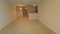 This single-floor space presents an open and functional layout with a warm, neutral color palette throughout. The main living area is finished with wall-to-wall beige carpeting, contributing to a cozy and understated ambiance. Smooth, cream-colored walls and white baseboards provide a clean aesthetic framework.

A half-wall partition separates the kitchen area from the living space, featuring what appears to be a granite or stone-topped counter, offering both visual separation and casual seating or serving potential. The kitchen, partially visible beyond the partition, includes standard cabinetry and appliances.

A pendant light fixture hangs above the kitchen or dining area, complementing the flush-mount ceiling light that illuminates the main living space. A floor-level air vent is visible along the wall, indicating a standard forced-air climate system.

A hallway extending from the main living area leads to additional rooms, with overhead lighting guiding the path. The corridor provides access to what appears to be closet space and further living areas. Overall, the space offers a straightforward, move-in-ready environment with consistent finishes throughout.
