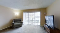 This single-floor residence offers a welcoming and practical living environment designed for everyday comfort. The living room serves as the focal point of the space, showcasing large sliding glass doors fitted with vertical blinds that allow for adjustable natural light and provide direct access to an outdoor area. The expansive glazing brings in a generous amount of daylight, creating an open and airy atmosphere throughout the room. Soft, wall-to-wall carpeting in a neutral tone adds warmth underfoot and complements the light-colored walls, which carry a subtle warm hue. A textured popcorn ceiling adds a classic residential character to the space. The layout accommodates comfortable seating and an entertainment setup, with ample floor space for furniture arrangement. Floor lamps positioned on either side of the room provide supplemental ambient lighting for evening use. The overall ambiance is calm and livable, with a straightforward design that prioritizes functionality and comfort. The connection to the outdoors through the sliding doors enhances the sense of space, making the living area feel larger and more connected to its surroundings.