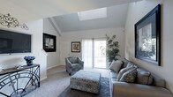 This two-floor space is highlighted by a striking living area that immediately draws attention with its vaulted ceiling and centrally placed skylight, which floods the room with abundant natural light. The angled ceiling adds a sense of height and architectural interest, creating an airy, open atmosphere. Soft neutral-toned carpet and crisp white walls contribute to a clean, modern aesthetic throughout.

The living area is thoughtfully arranged with a grey sectional sofa accented by patterned throw pillows, a coordinating patterned accent chair, and a decorative upholstered ottoman that serves as a functional centerpiece. A large area rug anchors the seating arrangement, adding texture and depth to the space. Framed artwork on the walls adds character without overwhelming the understated palette.

A full-length sliding glass door provides direct access to the outdoors and frames views of lush greenery, enhancing the connection between interior and exterior. A tall indoor plant beside the door reinforces this natural element. Staircase access is visible, indicating the multi-level layout of the home, with additional living space found on the second floor.