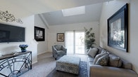 This two-floor space is highlighted by a striking living area that immediately draws attention with its vaulted ceiling and centrally placed skylight, which floods the room with abundant natural light. The angled ceiling adds a sense of height and architectural interest, creating an airy, open atmosphere. Soft neutral-toned carpet and crisp white walls contribute to a clean, modern aesthetic throughout.

The living area is thoughtfully arranged with a grey sectional sofa accented by patterned throw pillows, a coordinating patterned accent chair, and a decorative upholstered ottoman that serves as a functional centerpiece. A large area rug anchors the seating arrangement, adding texture and depth to the space. Framed artwork on the walls adds character without overwhelming the understated palette.

A full-length sliding glass door provides direct access to the outdoors and frames views of lush greenery, enhancing the connection between interior and exterior. A tall indoor plant beside the door reinforces this natural element. Staircase access is visible, indicating the multi-level layout of the home, with additional living space found on the second floor.