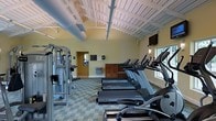 This single-story fitness center offers a thoughtfully organized workout space designed to accommodate a range of exercise needs. The room features an open layout with exposed white-painted beam ceilings that lend an airy, spacious feel, while visible ductwork adds an industrial touch overhead. The walls are finished in a warm yellow tone, contributing to an energizing atmosphere throughout.

The cardio area is prominently outfitted with a row of TRUE-brand treadmills aligned along the central floor space, complemented by elliptical machines positioned toward the windowed wall. Natural light filters in through a window on one side, brightening the workout area. A wall-mounted flat-screen television is installed at an elevated position, providing entertainment for cardio users.

Along the opposite wall, a multi-station strength training machine offers a variety of resistance-based exercise options. The flooring consists of a patterned carpet tile that provides cushioning underfoot. Additional amenities include a water fountain and a wooden storage cabinet near the far wall. A doorway leads to an adjoining room, suggesting further facilities are accessible within the space.