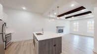 Located at 21 Forest Ridge Blvd in Mechanicville, NY, this single-floor home offers a thoughtfully designed open-concept layout with modern finishes throughout. The kitchen serves as the centerpiece of the main living area, featuring a large island with a white quartz countertop, an undermount sink, and dark wood-toned cabinetry. A trio of pendant lights hang above the island, complementing the recessed lighting overhead. Stainless steel appliances, white upper cabinetry, and light luxury vinyl plank flooring complete the kitchen's clean, contemporary aesthetic. The kitchen flows seamlessly into the spacious living room, which is distinguished by striking exposed dark wood ceiling beams and a built-in fireplace with a wood mantel. A window allows natural light to fill the room, enhancing the warm, inviting atmosphere. The bathrooms reflect the same refined style — the primary bath includes a double-sink vanity with quartz countertops, dark cabinetry with brushed gold hardware, large-format tile flooring, and a tiled shower. A secondary full bath features a tub-shower combo with clean white finishes. The bedrooms are carpeted for comfort and include wall-mounted climate control units. Throughout, the home maintains a bright, neutral palette with crisp white walls and modern trim details.