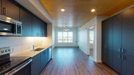 Located at 1090 NE 91st Ave in Hillsboro, Oregon, this thoughtfully designed single-floor residence combines modern finishes with functional living spaces. The kitchen is a standout feature, showcasing sleek dark navy cabinetry, light quartz countertops, a white subway tile backsplash, and a full suite of stainless steel appliances including a refrigerator, electric range, over-the-range microwave, and dishwasher. A distinctive warm wood-plank ceiling extends from the kitchen into the open living area, adding a natural, inviting contrast to the clean white walls and dark hardwood-style flooring beneath. The living area is generously proportioned, illuminated by a ceiling fan with integrated lighting and a large window that frames outdoor views and draws in abundant natural light. The bedroom continues the warm aesthetic with carpeted flooring, a ceiling fan, wood-plank ceiling detail, and a bright window overlooking the surrounding landscape. The bathroom is well-appointed with a soaking tub and shower combination, built-in wall shelving, a dark-toned vanity with an undermount sink, and hardwood-style flooring. The overall design balances contemporary style with comfortable, practical living throughout.
