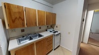 This single-floor unit offers a straightforward, functional layout across a compact but well-organized footprint. The living area is finished with wall-to-wall carpet in a neutral tan tone, providing a warm underfoot feel. The space is illuminated by a centrally mounted ceiling light and features baseboard heating along the perimeter walls. A wall-unit air conditioner is installed beneath a window fitted with blinds, offering both climate control and natural light. A mirrored sliding closet adds depth and storage to the bedroom area, which connects openly to the main living space.

The galley-style kitchen is efficiently arranged along one wall, featuring warm-toned wood cabinetry both above and below the counter. A double stainless steel sink, a white electric coil-top range, and a full-size white refrigerator provide all essential appliances. A tiled backsplash with accent details runs behind the countertop, and light tile flooring keeps the kitchen area easy to maintain.

The bathroom is finished in neutral tones with a marbled laminate vanity countertop, an integrated sink, and a large mirror with overhead lighting. A tub with a shower rod, a toilet, baseboard heating, and tile flooring complete the functional space. Overall, the unit presents a clean, no-frills living environment.
