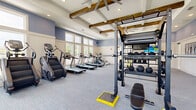Located at 4910 Samaritan Way in Jacksonville, FL, this fitness center offers a spacious, single-floor layout designed to support a full range of workout routines. The space is architecturally distinguished by a striking coffered ceiling accented with rustic exposed wood beams and oversized ceiling fans, creating a refined yet functional atmosphere. Soft blue and neutral wall tones complement the light gray carpeted flooring throughout.

Large windows line multiple walls, flooding the interior with natural light and offering views of the surrounding outdoor greenery — an inviting backdrop for any workout. Full-length mirrors are strategically positioned to enhance the sense of space and support form-focused training.

The cardio zone features a row of Spirit treadmills, stair climbers, and elliptical machines, all arranged to take advantage of the natural light and outdoor views. The strength training area is equally well-appointed, with a multi-functional power rack system, TRX suspension trainers, a full dumbbell rack, barbells, weight plates, medicine balls, and an adjustable bench. Additional cable and resistance machines round out the equipment offerings.

The open floor plan ensures ample circulation space between stations, making the facility comfortable for multiple users simultaneously. Double glass doors connect to adjacent amenity spaces, adding convenience and accessibility to this thoughtfully designed fitness facility.