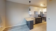 Located at 2333 Jackson St NE in the vibrant Northeast Minneapolis neighborhood, this single-floor unit showcases a thoughtfully designed, contemporary interior with a clean, modern aesthetic. Light gray wood-plank flooring runs throughout the space, creating a cohesive and polished look from room to room.

The kitchen is a standout feature, offering a functional galley-style layout with dark espresso lower cabinetry contrasted by crisp white upper cabinets. Stainless steel appliances — including a range, microwave, and dishwasher — complement the neutral countertops, while a peninsula with an integrated sink provides additional prep space and casual seating potential. A stylish pendant light anchors the kitchen area, adding a modern touch.

The living area is open and versatile, benefiting from generous natural light streaming through large windows that frame views of the surrounding residential neighborhood and mature tree canopy. The neutral warm-toned walls provide a welcoming backdrop throughout.

The bathroom maintains the same modern sensibility, featuring a dark-toned vanity with ample storage, a white integrated sink, and tile-style flooring that mirrors the rest of the unit. The layout is efficient and well-appointed with accessibility-friendly grab bar hardware.

Overall, this unit presents a well-executed modern design with quality finishes, an abundance of natural light, and a practical, open layout.