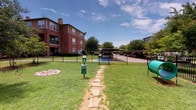 Located at 1420 W McDermott Dr in Allen, Texas, this outdoor community space offers a thoughtfully designed array of amenities spread across generously landscaped grounds. The property is characterized by mature, towering trees that provide ample shade and a lush, park-like atmosphere throughout. Winding concrete pathways connect the various areas of the community, flanked by mulched tree beds and well-kept green lawns.

A dedicated children's playground features a multi-level play structure with dual slides, climbing elements, and a wood chip safety surface, set against a backdrop of verdant trees and brick residential buildings. Nearby, a full-size basketball court offers an open, well-marked surface surrounded by neatly arranged evergreen trees under an open Texas sky.

The grounds also include a dog-friendly area, complete with a stone-paved walking path, waste stations, and play equipment for pets, enclosed by black iron fencing. Shaded picnic areas with wooden tables and benches are nestled beneath large canopy trees, providing inviting spots for outdoor gathering. The overall ambiance is green, open, and community-focused, making this property an appealing and well-equipped outdoor environment for residents of all ages.