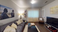 This single-floor unit is designed with an open-concept layout that seamlessly connects the kitchen and living area, creating a cohesive and functional living space. The kitchen features dark cabinetry paired with granite-style countertops and a peninsula that doubles as a breakfast bar, complete with industrial-style stools. A pendant light hangs above the peninsula, providing focused task lighting, while black appliances—including a refrigerator, microwave, and dishwasher—complement the cabinetry. Wood-style flooring runs throughout the unit, adding warmth and visual continuity.

The living area is positioned adjacent to the kitchen and receives natural light through a large window dressed with sheer curtains. The space comfortably accommodates a sofa, coffee table, and accent seating, offering a relaxed yet organized arrangement.

The bedroom is a well-proportioned private retreat, featuring a window with sheer curtains, a dedicated desk area suited for work or study, and ample room for a full-sized bed. The bathroom includes a granite-topped vanity with dark cabinetry, a toilet, and a full tub-shower combination, maintaining the consistent design aesthetic found throughout the unit. Overall, the neutral gray walls and wood-style flooring provide a clean, modern backdrop across all rooms.