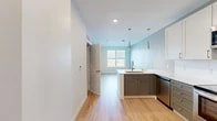 Located at 215 E State St in downtown Ithaca, NY, this one-bedroom, one-bathroom unit offers a thoughtfully designed layout that maximizes its compact footprint. Light-toned hardwood flooring runs continuously throughout, creating a cohesive and clean aesthetic across all rooms.

The open kitchen and living area serves as the heart of the unit. The kitchen is well-appointed with two-tone cabinetry — grey shaker-style lower cabinets paired with white upper cabinets — complemented by white countertops and a white tile backsplash. A full suite of stainless steel appliances includes a refrigerator, electric range, over-the-range microwave, and dishwasher. A peninsula with a sink and pendant lighting above adds both functionality and visual interest, while soft blue-grey accent walls lend a modern touch.

The bedroom is a quiet, compact space featuring a large window with horizontal blinds that frames elevated views of downtown Ithaca's skyline and surrounding greenery, filling the room with natural light.

The bathroom features a glass-enclosed walk-in shower with white subway tile walls, offering a clean, contemporary finish. A dedicated closet and entry area complete the unit's efficient floor plan.