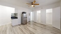 Located at 4451 Lockwood Ave in the East Hollywood neighborhood of Los Angeles, this single-floor studio offers a practical and efficiently designed layout. The main living area doubles as the kitchen and living room, finished with light wood-style vinyl flooring that runs throughout and crisp white walls that lend a clean, bright atmosphere. A ceiling fan centered overhead provides both airflow and ambient lighting, while a sizable window along the exterior wall allows natural light to filter in and offers a view of the surrounding greenery. The kitchen area is positioned along one wall and features dark countertops, mosaic tile backsplash, a stainless steel sink, and stainless steel appliances including a refrigerator and microwave, maximizing functionality within a compact footprint. A dedicated closet provides essential storage space. The bathroom is finished with white tile throughout and includes a full bathtub and shower combo equipped with a modern square rainfall showerhead and chrome fixtures, a pedestal-style sink, and a toilet. The overall design emphasizes clean lines and a neutral palette, creating a cohesive and low-maintenance living environment well-suited for those seeking practical urban living in Los Angeles.