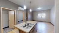 Located at 1307 Hayes St in La Crosse, Wisconsin, this single-floor unit offers a thoughtfully designed layout with a clean, modern aesthetic. The open-concept kitchen and living area serves as the heart of the space, featuring light hardwood-style flooring, warm wood cabinetry extending to the upper walls, black appliances including a gas range and full-size refrigerator, and a central island with a double stainless steel sink accented by pendant lighting overhead. A wall-mounted mini-split unit provides efficient climate control, complemented by baseboard heating throughout. The living area benefits from natural light through a vertical-blind window, creating a bright and welcoming atmosphere. The bedroom is generously proportioned with plush carpeting, a large window, and baseboard heating, offering a comfortable and quiet retreat. The bathroom is designed with accessibility in mind, featuring grab bars, a wide layout, a dark wood vanity with an integrated tall storage cabinet, a well-lit mirror, and neutral tile-style flooring. The unit's overall palette of gray, beige, and warm wood tones creates a cohesive and contemporary feel, making this a functional and comfortable living space in La Crosse.