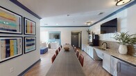 Located at 2200 House Creek Trail in Raleigh, NC, this single-floor community amenity space is thoughtfully designed to serve as a versatile gathering hub. The layout is divided into several interconnected zones that flow seamlessly into one another, creating a cohesive yet functionally distinct environment.

The lounge area is a standout feature, characterized by tray ceilings painted in a bold navy blue that adds depth and visual interest. Comfortable seating arrangements include upholstered armchairs with nailhead trim, modular blue ottoman cubes, and neutral sofas accented with teal and orange throw pillows. Patterned area rugs and warm hardwood-style flooring tie the space together, while an array of framed artwork adorns the walls. Large windows dressed with floor-length curtains flood the room with natural light and offer views of the outdoor pool area.

A generous dining zone accommodates a long communal table flanked by leather chairs, ideal for group gatherings or meetings. Colorful gallery-style artwork lines the walls, reinforcing the space's creative aesthetic.

The kitchenette is well-appointed with granite countertops, a tile backsplash, white cabinetry, a stainless steel under-counter refrigerator, a sink, and open shelving. A wall-mounted television above the counter adds convenience. Restroom facilities are also accessible from this area, making the space fully self-contained for events and everyday use.