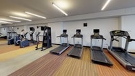 This fitness center is a comprehensively outfitted facility designed to support a full range of workout routines. The space is organized into distinct functional zones, providing an efficient and comfortable environment for exercise. The cardio area is lined with multiple treadmills, each equipped with individual screen displays, alongside elliptical machines, offering ample options for aerobic training. Complementing these are strength training stations, including cable and multi-function weight machines, as well as a dedicated free weights section featuring a tiered dumbbell rack with a full set of weights and an adjustable bench. A row of stair-climber machines further expands the cardio offerings. The flooring combines warm wood-tone panels with striped carpet sections, visually delineating different workout zones throughout the room. Structural concrete columns punctuate the interior, adding an industrial character to the design. Large floor-to-ceiling windows along one wall flood the space with natural light and offer views of a landscaped outdoor courtyard with mature trees, creating an inviting atmosphere. The ceiling features linear fluorescent lighting that ensures consistent, bright illumination throughout. Full-length mirrors in the strength training area enhance spatial openness and allow users to monitor their form. A water fountain is also conveniently accessible within the space.
