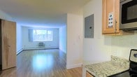 This single-floor space offers a clean, functional layout that connects the kitchen and living areas in an open, flowing arrangement. The living area is finished with warm hardwood flooring that reflects natural light streaming in through a large window, which frames a view of outdoor greenery. A wall-mounted HVAC unit and baseboard heating provide year-round climate control, while white walls throughout contribute to a bright, neutral atmosphere.

The kitchen is situated adjacent to the main living space and features granite countertops with a speckled finish and light wood cabinetry that adds warmth to the area. A structural dividing wall separates the two zones and houses an accessible electrical panel, a practical detail that keeps utilities within easy reach.

The overall design is straightforward and functional, with consistent hardwood flooring tying the spaces together seamlessly. The large window in the living area serves as a focal point, drawing in natural light and creating a sense of openness. The space is well-suited for those seeking a simple, low-maintenance layout with key amenities integrated efficiently throughout.