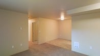 This single-floor residential unit offers a straightforward and practical layout designed for comfortable everyday living. The main living area is finished with wall-to-wall carpeting in a neutral beige tone, providing a warm and cohesive feel throughout the space. Walls are painted in a soft, light neutral color that helps maximize the brightness of the interior, complemented by a flush-mount ceiling light fixture that provides even, ambient illumination. A floor-level HVAC vent is integrated into the baseboard, ensuring efficient climate control throughout the unit. The living area flows naturally toward a hallway that connects to additional rooms and a bathroom, which features a glass-door shower enclosure visible from the main space. Subtle architectural details, including a soffit element near the entry area, add visual structure to the open layout. The unit's simple, clean design makes it highly adaptable for a variety of furnishing arrangements, offering a blank canvas for personalization. Overall, the space presents a functional and unpretentious residential environment with consistent finishes and a calm, neutral ambiance throughout.