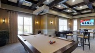This sophisticated entertainment space is thoughtfully designed to balance style with leisure. The room's most striking architectural feature is its dark-stained coffered ceiling, which adds a sense of depth and refined character to the overall ambiance. Recessed lighting embedded within the ceiling grid provides warm, even illumination throughout the space.

At the heart of the room sits a full-size billiards table with a cream-toned felt surface, serving as the central focal point for recreation and social gatherings. Alongside it, a dark grand piano occupies a dedicated corner, lending a cultivated, upscale atmosphere that elevates the room beyond a typical game room.

Large windows along the walls invite abundant natural light and offer pleasant views of the surrounding greenery, creating an airy and open feel. Elegant drapes with a geometric pattern frame each window, adding a layer of texture and warmth. Wall-mounted sconces positioned between the windows provide soft accent lighting that complements the room's warm, golden wall tones.

A chair rail and wainscoting detail along the lower walls contribute a classic architectural touch, reinforcing the room's polished, upscale character. Overall, this space combines functional entertainment amenities with refined design elements, making it an inviting and distinguished gathering area.