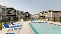 This outdoor amenity space centers around a generously sized, crystal-clear swimming pool with a clean tile border and built-in steps for easy entry. The surrounding pool deck provides ample space for relaxation and social gathering, furnished with a mix of blue and white lounge chairs and Adirondack-style seating arranged around small side tables — ideal for unwinding in the open air. The area is fully enclosed by a black wrought iron-style fence with a gated entry, providing both security and an unobstructed view of the open landscape beyond. A white utility enclosure is situated along one side of the deck, keeping the space tidy and functional. The backdrop features the multi-story residential buildings of the surrounding community, which are finished in neutral gray siding with covered balconies and pitched rooflines, lending the pool area a welcoming, neighborhood feel. Manicured green lawn connects the pool enclosure to the adjacent buildings, enhancing the cohesive outdoor environment. The open sky above allows for abundant natural light throughout the day, making this pool deck a bright and inviting retreat for residents seeking outdoor recreation and relaxation.