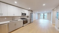 Located at 105 Washington St in Boston's Brighton neighborhood, this modern unit offers five bedrooms and two well-appointed bathrooms across a single thoughtfully designed floor. Light-toned hardwood flooring flows throughout, lending a clean, cohesive aesthetic to every room. The open-concept kitchen and living area serves as the heart of the unit, featuring a sleek galley-style kitchen outfitted with white upper cabinetry, two-tone gray lower cabinets, a subway tile backsplash, and a full suite of stainless steel appliances including a gas range, microwave, dishwasher, and French door refrigerator. Recessed lighting illuminates the space, while a warm wood-toned entry door adds a refined contrast. The living area extends toward floor-to-ceiling sliding glass doors that open to a private balcony, filling the room with natural light and urban views. The five bedrooms are well-sized, with the primary bedroom offering generous proportions and ample space. The two bathrooms are distinctly styled: one features a clean white tub-shower combination, while the other is elevated with a double-sink vanity and a glass-enclosed walk-in shower adorned with striking marble-look tile. Two hallways connect the layout efficiently, complemented by a dedicated closet for additional storage.