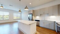 This well-designed single-floor space combines modern aesthetics with functional living. The open-concept kitchen is a standout feature, showcasing greige shaker-style cabinetry extending to the ceiling, a classic white subway tile backsplash, and a full suite of stainless steel appliances including a French door refrigerator, dishwasher, microwave, and range. A large central island with a clean white countertop provides additional prep space and casual seating, while two polished industrial-style pendant lights hang overhead. Warm, light-toned hardwood-style flooring flows throughout the main living areas, complementing the bright, neutral color palette. The living area is generously proportioned with large windows dressed in white blinds, a ceiling fan, and ample natural light. The bedroom continues the clean, airy feel with plush wall-to-wall carpeting, a ceiling fan, and a wide window offering views of surrounding greenery. The bathroom features a long vanity with a white countertop and grey cabinetry, a tiled shower-tub combination, and hardwood-style flooring. Throughout the space, crisp white walls, recessed lighting, and consistent modern finishes create a cohesive and inviting atmosphere.