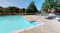 This amenity space is composed of two primary areas: a fully equipped fitness center and an outdoor swimming pool, together forming a comprehensive recreational facility. The fitness center is accessed through double glass doors clearly marked "Fitness Center," lending a clean, professional entry point. Inside, the space is well-lit by a combination of recessed ceiling lights and large windows with roller shades, which frame direct views of the outdoor pool. The gym floor features a diverse array of cardio equipment, including multiple treadmills, elliptical trainers, and stationary bikes — both upright and recumbent — as well as strength-training machines from brands such as Hoist. A freestanding dumbbell rack adds to the free weights selection. Wall-mounted televisions are positioned for convenient viewing during workouts, and the warm accent walls give the space an inviting atmosphere. The outdoor pool area features a generously sized swimming pool with clearly marked depth indicators, surrounded by a wide concrete deck with lounge chairs available for relaxation. The pool is enclosed by a metal fence and set against a lush, tree-lined backdrop, providing a pleasant and shaded environment. Together, these amenities offer a well-rounded and functional space suited for fitness and leisure.