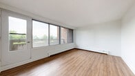 Located at Woodridge Crescent in Ottawa, Ontario, this one-bedroom unit offers a thoughtfully organized single-floor layout comprising a living room, bedroom, and kitchen. The living room is a standout feature, characterized by a wide bank of sliding windows that flood the space with natural light and frame views of the surrounding neighbourhood, including glimpses of greenery and nearby buildings. Warm wood-look laminate flooring runs throughout the living and sleeping areas, lending a cohesive and inviting feel, while baseboard heating units are positioned along the exterior walls. The bedroom is a comfortable, well-proportioned room featuring a sliding-door closet and a window that continues the theme of natural light and outdoor views. A clean white colour palette across all rooms enhances the sense of openness. The kitchen is finished with modern white cabinetry, dark stone-effect countertops, a white tile backsplash, and light grey tile flooring. A stainless steel electric range with an overhead ventilation hood serves as the kitchen's focal point, complemented by ample upper and lower cabinet storage. The overall design is clean and functional, with a neutral aesthetic that provides a versatile foundation throughout.