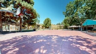 Located at 9300 Montana Ave in El Paso, Texas, this outdoor recreational area offers a well-equipped sporting facility set against a backdrop of mature trees and open blue skies. The centerpiece of the space is a full-sized basketball court with a painted surface, complete with regulation hoops at both ends and clearly marked court lines. Adjacent to the basketball court is a tennis court, enclosed by a chain-link fence that defines the playing area and provides wind protection. A notable feature of the complex is a shaded gathering area covered by a large green sail-shade canopy, supported by sturdy posts and surrounded by raised seating or planter borders, offering a comfortable spot for spectators or community gatherings. The courts are illuminated by tall pole-mounted light fixtures, making the space functional during evening hours. Lush, mature trees — including pines and deciduous varieties — frame the entire area, providing natural shade and greenery that contrast with the warm desert surroundings. The neighboring multi-story residential buildings, finished in a sandy beige stucco, are visible from the courts, situating the recreational space firmly within a larger residential community. Well-maintained landscaping with trimmed shrubs and grassy areas completes the grounds.