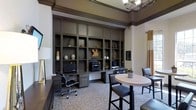 This business center offers a well-appointed, single-floor workspace designed to meet a range of professional needs. The room is anchored by an expansive wall of floor-to-ceiling built-in shelving finished in a rich, dark tone, providing generous storage and display space alongside integrated workstations equipped with desktop computers and a shared printer. The cabinetry's detailed millwork and column-style pilasters lend a sense of architectural sophistication to the space. Crown molding with a contrasting dark trim band lines the upper walls, adding a layer of classic elegance. Elevated ceilings contribute to an open, airy feel, while a decorative chandelier provides warm overhead lighting. A round high-top table with sleek bar-height chairs occupies the center of the room, offering an informal setting for collaboration or independent work. Neutral wall tones, warm drapery accents, and tasteful decorative objects throughout create a refined yet functional ambiance. The overall design balances professional utility with a polished aesthetic, making this business center a welcoming and efficient environment for work and productivity.