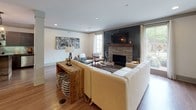 This single-floor space offers a thoughtfully designed open layout that seamlessly connects the living area, transitional spaces, and kitchen into one cohesive environment. The living room serves as the centerpiece of the home, anchored by a striking floor-to-ceiling stone fireplace clad in dark wood-toned tile and fitted with a rustic wood mantle. A wall-mounted television sits above the fireplace, flanked by large windows that invite abundant natural light and offer views of the surrounding greenery. Rich, dark hardwood flooring in the living area gives way to lighter wood-toned flooring throughout the rest of the space, adding visual warmth and depth. Recessed lighting throughout keeps the atmosphere bright and modern. The open-concept kitchen features dark cabinetry, a green glass tile backsplash, stainless steel appliances, and a rustic bar-height island with seating — ideal for casual dining or entertaining. Industrial-style pendant lights hang above the island, adding character and dimension. Adjacent flex areas provide versatile space for additional seating or display, accented by bold typographic wall art. Crown molding and clean architectural lines contribute to the polished yet approachable character of the overall space.