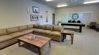 This single-floor space is designed to balance comfort and functionality, featuring a well-appointed lounge area that serves as a central gathering space. The lounge is anchored by a generously sized tan leather sectional sofa, complemented by rustic reclaimed wood coffee and side tables that add a warm, earthy character to the room. Wildlife-themed framed artwork, including images of giraffes, zebras, and horses, adorns the light gray walls, lending the space a distinctive, curated aesthetic.

Overhead, flush-mounted fluorescent lighting provides consistent illumination throughout. The neutral-toned carpeted flooring ties the space together cohesively. A decorative potted plant near the back wall introduces a touch of greenery, enhancing the welcoming atmosphere.

Adjacent to the lounge, a windowed meeting or conference room is visible, featuring a large wall-mounted clock as a focal point and several chairs arranged around what appears to be a work surface. The glass partition allows visual connectivity between the two areas while maintaining a sense of separation. Overall, the space presents a professional yet approachable environment suited for both relaxed interaction and focused work.
