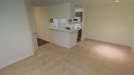 This single-floor residence offers a straightforward and functional layout designed for comfortable everyday living. The open-concept living area is finished with plush wall-to-wall carpeting in a neutral beige tone, creating a warm and cohesive feel throughout the space. Light-colored walls contribute to a bright, airy atmosphere.

The kitchen is positioned at the rear of the main living area and is defined by a raised pass-through breakfast bar that serves as a natural divider between the cooking and living spaces. The kitchen features white appliances, including a refrigerator, electric range, and overhead microwave, complemented by medium-toned wood cabinetry that provides ample storage. The kitchen floor transitions to a dark hardwood or wood-look surface, offering a practical and easy-to-clean contrast to the carpeted living area.

The overall design prioritizes an open flow between the kitchen and living room, making the space feel connected and accessible. Neutral finishes throughout — from the walls to the flooring — provide a versatile backdrop that accommodates a variety of décor styles. The layout is efficient and well-organized, making thoughtful use of the available footprint across its single level.