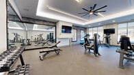 This modern fitness center offers a generously sized, single-floor layout designed to accommodate a full range of exercise activities. The space is defined by its striking architectural details, including tray ceilings accented with recessed LED strip lighting and large industrial-style ceiling fans that ensure consistent air circulation throughout the room. Durable speckled flooring covers the entire space, providing a practical and clean surface suited for high-traffic fitness use.

The facility is outfitted with a comprehensive selection of equipment, including treadmills, cable machines, selectorized strength machines, adjustable benches, a multi-station cable system, and a dedicated free weight area featuring a tiered dumbbell rack. A full-length wall mirror on one side of the room visually expands the space and allows users to monitor their form during workouts. Mounted flat-screen televisions are positioned throughout for entertainment and instruction.

Textured stone accent walls add warmth and visual interest, complemented by framed fitness-themed artwork that reinforces the athletic atmosphere. Large floor-to-ceiling windows and a sliding glass door along the perimeter flood the space with natural light and offer views of the surrounding outdoor area. The overall ambiance is clean, modern, and purposefully designed to support a motivating and functional fitness environment.
