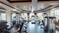 This fitness center is designed with both functionality and aesthetic refinement in mind. The space is defined by its striking coffered ceilings with dark wood trim detailing, recessed lighting, and crisp white walls that lend a polished, upscale atmosphere. Large multi-pane windows wrap around the perimeter of the room, flooding the interior with natural light and offering picturesque views of surrounding tropical greenery and palm trees, creating an inviting environment for exercise.

The cardio zone is well-equipped with treadmills, elliptical trainers, stationary bikes, and a recumbent cycle, all positioned facing the windows to take full advantage of the outdoor scenery. Wall-mounted flat-screen televisions are also positioned throughout this area for entertainment during workouts. The strength training section provides a thorough selection of selectorized cable machines and resistance equipment suited for a full-body workout. A dedicated free weights area features a complete dumbbell rack with an arched mirror backdrop, adding depth to the space.

Dark rubber flooring throughout provides durability and safety underfoot. Rich dark wood entry doors and arched mirror accents complement the refined architectural details. The overall ambiance is bright, spacious, and well-organized, offering a premium fitness experience.