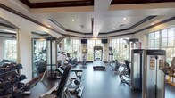 This fitness center is designed with both functionality and aesthetic refinement in mind. The space is defined by its striking coffered ceilings with dark wood trim detailing, recessed lighting, and crisp white walls that lend a polished, upscale atmosphere. Large multi-pane windows wrap around the perimeter of the room, flooding the interior with natural light and offering picturesque views of surrounding tropical greenery and palm trees, creating an inviting environment for exercise.

The cardio zone is well-equipped with treadmills, elliptical trainers, stationary bikes, and a recumbent cycle, all positioned facing the windows to take full advantage of the outdoor scenery. Wall-mounted flat-screen televisions are also positioned throughout this area for entertainment during workouts. The strength training section provides a thorough selection of selectorized cable machines and resistance equipment suited for a full-body workout. A dedicated free weights area features a complete dumbbell rack with an arched mirror backdrop, adding depth to the space.

Dark rubber flooring throughout provides durability and safety underfoot. Rich dark wood entry doors and arched mirror accents complement the refined architectural details. The overall ambiance is bright, spacious, and well-organized, offering a premium fitness experience.