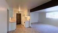 This single-floor space offers a practical and functional layout centered around an open-concept design that connects the kitchen and living area. The kitchen is equipped with white cabinetry, a white electric range, a refrigerator, and an under-cabinet range hood, providing all the essentials for everyday cooking. A tile floor runs through the kitchen, offering easy maintenance, while a dark exposed beam serves as a defining architectural element that visually separates the kitchen from the adjoining living area. The living space transitions to plush carpeting, creating a warm and comfortable atmosphere. White walls throughout keep the interior feeling bright and open. A louvered dark wood door adds a rustic contrast to the otherwise clean, neutral palette and leads to an additional room or utility area. A ceiling-mounted light fixture provides overhead illumination in the kitchen, complementing the under-cabinet lighting. The layout is straightforward and efficient, making the most of the available floor space on a single level. Overall, the space presents a simple, no-frills environment well-suited for comfortable everyday living.