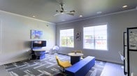 This single-floor space is a thoughtfully designed communal lounge that blends modern aesthetics with functional comfort. The room is anchored by a bold color palette, featuring striking blue and yellow furnishings that create a lively and energetic atmosphere. A plush blue bench and yellow accent chairs are arranged around a round central table, offering a casual seating area ideal for conversation or collaboration. A dedicated workstation along the wall includes a flat-screen television, a sleek desk, and designer wire-frame chairs, providing a functional space for work or entertainment. Large windows line two walls, flooding the interior with abundant natural light while offering views of the surrounding exterior. The ceiling features crown molding, recessed lighting, and a contemporary ceiling fan, contributing to the room's polished yet relaxed feel. Patterned carpet tiles in muted tones ground the space and complement the vibrant furnishings. Colorful wall art adds creative flair throughout, reinforcing the space's modern and welcoming character. Overall, the lounge offers a well-balanced environment that is both visually dynamic and practically equipped for a variety of uses.