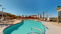 Located in Las Cruces, New Mexico, this outdoor amenity area offers a resort-style retreat designed to complement the sunny Southwest climate. At the heart of the space is a generously sized freeform swimming pool with a smooth curved shape, accessible via multiple entry points with handrail-equipped steps. Adjacent to the main pool sits a dedicated hot tub, providing a relaxing counterpart to the larger swimming area. The expansive concrete sun deck surrounds both water features and is outfitted with rows of cushioned lounge chairs, perfect for soaking in the abundant New Mexico sunshine. A wooden pergola anchors one end of the deck, offering a shaded gathering spot beneath the open sky. Tall palm trees and slender Italian cypress trees are strategically placed throughout the area, adding vertical interest and a touch of desert elegance. The entire pool area is enclosed by a decorative metal fence, ensuring privacy and security. Umbrella-shaded seating stations provide additional relief from the sun, while ample deck space allows for comfortable circulation. The surrounding multi-story residential buildings, with their warm earth-toned and contemporary façades, frame the courtyard and contribute to a cohesive, community-centered atmosphere ideal for outdoor relaxation and recreation.
