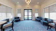 Located at 1 Patriots Place in Millbury, Massachusetts, this single-story space presents a polished and well-appointed interior designed with both aesthetic appeal and practicality in mind. The layout flows seamlessly from a grand central corridor to distinct gathering areas, each thoughtfully curated with complementary finishes throughout.

The main hallway makes an immediate impression with rich hardwood flooring, deep gray walls, crisp white crown molding, and decorative column details. Pendant lantern fixtures suspended from a tray ceiling cast a warm glow along the corridor, which leads toward a glass-paneled entry door that draws in natural light from outside.

The lounge area is a standout feature of the space, showcasing a distinctive blue-and-white patterned area rug anchoring the room. Tufted navy blue accent chairs and a multi-globe chandelier contribute to the sophisticated ambiance. French doors open directly to an outdoor area, further enhancing the sense of openness.

An adjacent dining nook offers a round bistro-style table paired with dark upholstered chairs beneath a dome pendant light, with roller shades on surrounding windows providing adjustable natural light. The combination of hardwood flooring, neutral wall tones, and shiplap ceiling details ties the entire space together in a cohesive, upscale aesthetic.