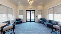 Located at 1 Patriots Place in Millbury, Massachusetts, this single-story space presents a polished and well-appointed interior designed with both aesthetic appeal and practicality in mind. The layout flows seamlessly from a grand central corridor to distinct gathering areas, each thoughtfully curated with complementary finishes throughout.

The main hallway makes an immediate impression with rich hardwood flooring, deep gray walls, crisp white crown molding, and decorative column details. Pendant lantern fixtures suspended from a tray ceiling cast a warm glow along the corridor, which leads toward a glass-paneled entry door that draws in natural light from outside.

The lounge area is a standout feature of the space, showcasing a distinctive blue-and-white patterned area rug anchoring the room. Tufted navy blue accent chairs and a multi-globe chandelier contribute to the sophisticated ambiance. French doors open directly to an outdoor area, further enhancing the sense of openness.

An adjacent dining nook offers a round bistro-style table paired with dark upholstered chairs beneath a dome pendant light, with roller shades on surrounding windows providing adjustable natural light. The combination of hardwood flooring, neutral wall tones, and shiplap ceiling details ties the entire space together in a cohesive, upscale aesthetic.