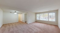 This single-floor unit offers a practical and functional layout designed for comfortable everyday living. The expansive main living area is finished with wall-to-wall neutral carpeting and features a ceiling fan with light fixture. Large windows along the front wall allow generous natural light to fill the space, providing views of the surrounding outdoor environment. A separate but open adjoining area extends the living space further, creating a versatile zone suitable for dining or additional seating arrangements.

The kitchen is equipped with white cabinetry, a gas range, a double-basin sink set into laminate countertops, and a dishwasher. A dedicated walk-in pantry adjacent to the kitchen provides ample dry storage. The kitchen floor features hardwood-style flooring, distinguishing it from the carpeted living areas.

The bedrooms are generously sized, each with neutral carpet, adequate natural light from windows, and ceiling-mounted light fixtures. One bedroom includes a sliding-door closet with built-in shelving and hanging rods, while another features direct access to an adjoining room, offering added flexibility.

The bathroom presents a clean, all-white aesthetic with tile walls, a single-sink vanity with under-cabinet storage, a mirrored medicine cabinet, and a full tub and shower combination. The mosaic tile flooring adds a subtle decorative touch to the otherwise streamlined space.
