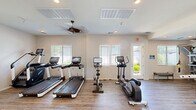 This fitness facility in Mesa, Arizona offers a comprehensive exercise space designed to accommodate a wide range of workout styles and fitness goals. The open, single-floor layout provides ample room for movement and equipment, all finished with durable wood-look flooring that adds warmth to the functional environment.

The cardio zone is well-appointed with multiple treadmills featuring interactive display screens, a stationary cycling bike, an elliptical trainer, and a stair climber — catering to various cardiovascular preferences. Ceiling fans circulate air throughout the space, enhancing comfort during intense workouts.

The strength training area is equally impressive, housing a multi-station cable machine, a selection of selectorized weight machines with padded blue seats, a power rack, and an adjustable workout bench. A well-stocked free weights section includes a full rack of dumbbells, kettlebells, and medicine balls to support functional and resistance training routines. TRX suspension straps are also available for bodyweight training.

Natural light pours in through multiple windows fitted with horizontal blinds, creating a bright and energizing atmosphere. Recessed lighting supplements the natural light for consistent illumination throughout the day. The crisp white walls and clean design aesthetic contribute to a welcoming, professional environment suited for daily fitness routines.