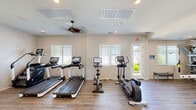 This fitness facility in Mesa, Arizona offers a comprehensive exercise space designed to accommodate a wide range of workout styles and fitness goals. The open, single-floor layout provides ample room for movement and equipment, all finished with durable wood-look flooring that adds warmth to the functional environment.

The cardio zone is well-appointed with multiple treadmills featuring interactive display screens, a stationary cycling bike, an elliptical trainer, and a stair climber — catering to various cardiovascular preferences. Ceiling fans circulate air throughout the space, enhancing comfort during intense workouts.

The strength training area is equally impressive, housing a multi-station cable machine, a selection of selectorized weight machines with padded blue seats, a power rack, and an adjustable workout bench. A well-stocked free weights section includes a full rack of dumbbells, kettlebells, and medicine balls to support functional and resistance training routines. TRX suspension straps are also available for bodyweight training.

Natural light pours in through multiple windows fitted with horizontal blinds, creating a bright and energizing atmosphere. Recessed lighting supplements the natural light for consistent illumination throughout the day. The crisp white walls and clean design aesthetic contribute to a welcoming, professional environment suited for daily fitness routines.