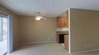 This single-floor space offers a straightforward and functional layout designed for comfortable everyday living. The main living area is finished with plush carpeting in a warm neutral tone, creating a cozy atmosphere throughout. A ceiling fan with an integrated light fixture is centrally mounted beneath a textured ceiling, providing both air circulation and ambient lighting to the open room.

The kitchen is seamlessly integrated into the living space, separated by a partial wall that creates a natural visual boundary while maintaining an open feel. The kitchen features warm honey-toned wood cabinetry both above and below the countertops, offering generous storage space. The countertops display a granite or stone-patterned surface, complementing the cabinetry tones. A stainless steel sink is positioned along the counter, and a black dishwasher is installed beneath the counter, adding to the kitchen's practicality. The kitchen floor transitions to a lighter tile, distinguishing it from the carpeted living area.

The walls throughout are painted in a consistent warm beige tone, contributing to a cohesive and inviting ambiance. White baseboards provide a clean contrast along the floors. Overall, the space presents a tidy, move-in ready environment with a practical and unified design aesthetic.