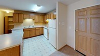 This single-floor unit offers a practical and comfortable layout designed for everyday living. The open-concept living area flows seamlessly into the kitchen, which is outfitted with warm oak cabinetry, white appliances including a range and refrigerator, a peninsula with countertop seating, and a patterned vinyl floor. A built-in shelving nook adjacent to the kitchen provides convenient storage space. Two carpeted bedrooms each feature neutral walls, baseboard heating, and windows fitted with blinds that bring in natural light. The textured ceilings are consistent throughout the space, lending a cohesive look to each room. Two full bathrooms serve the unit: one includes a single-sink vanity with oak cabinetry, a toilet, and a standalone shower, while the second features a combination tub and shower, a matching oak vanity, and light tile flooring. Both bathrooms are finished with white countertops and chrome fixtures. The overall palette of the space relies on warm beige and off-white tones, creating a clean and understated ambiance. Baseboard heating is present in multiple rooms, and the layout, as seen in the dollhouse view, reflects a well-organized floor plan with clear separation between living, sleeping, and utility areas.